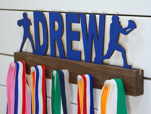 Our personalized football medal holder is a unique gift for the football player in your life. This display will be the perfect way for him to show off all of his well-deserved awards. This design comes in a variety of colors, or you can pick from our other choices of sports or phrases so we can customize it just the way you want!