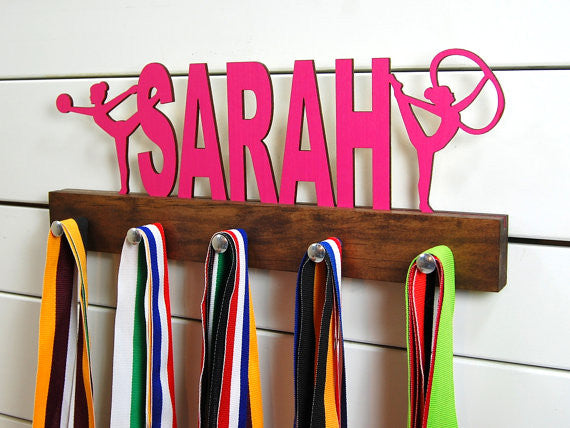  Our personalized rhythmic gymnastics medal holder is a unique gift for the gymnast in your life. This display will be the perfect way for her to show off all of her well-deserved awards. This design comes in a variety of colors, or you can pick from our other choices of sports or phrases so we can customize it just the way you want!